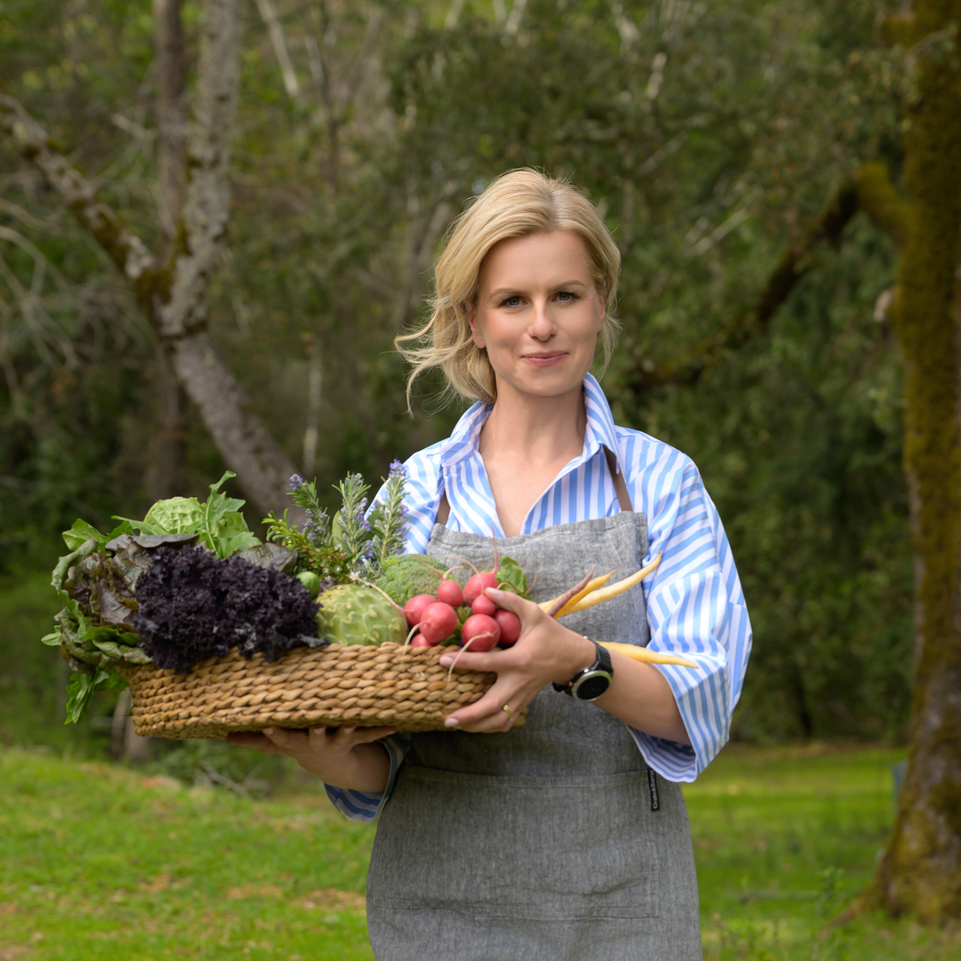Woman in a striped shirt and apron holding a basket filled with various fresh vegetables outdoors during her wellness coach personal branding session