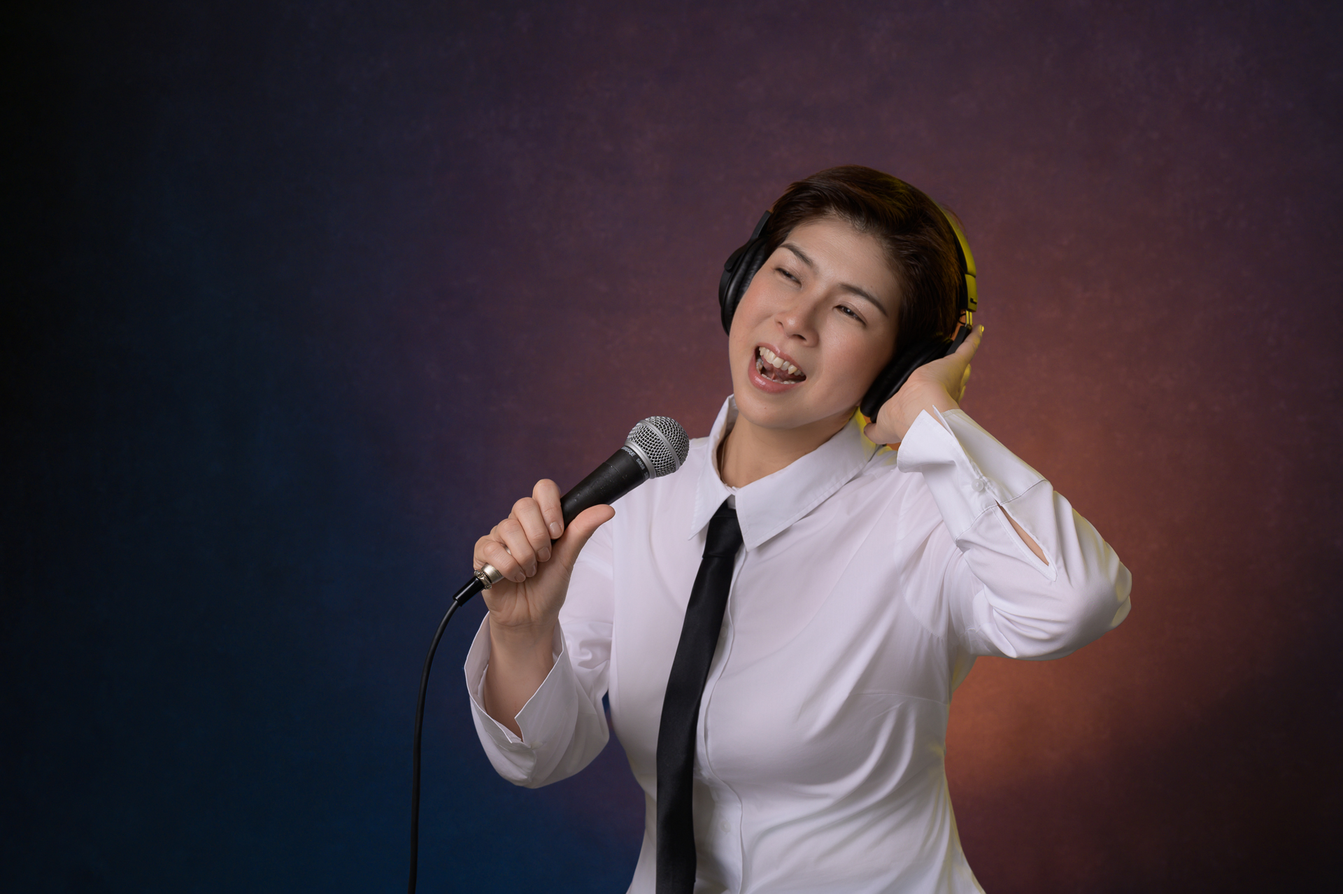 Woman wearing headphones and a white shirt singing into a microphone against a gradient background.
