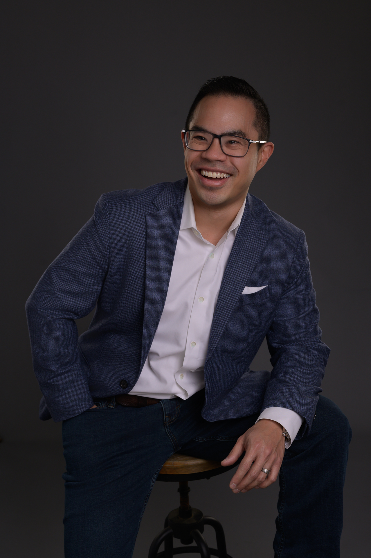 Thought Leader Branding Photos of a man wearing a blue blazer and white shirt, smiling while sitting on a stool against a dark background.