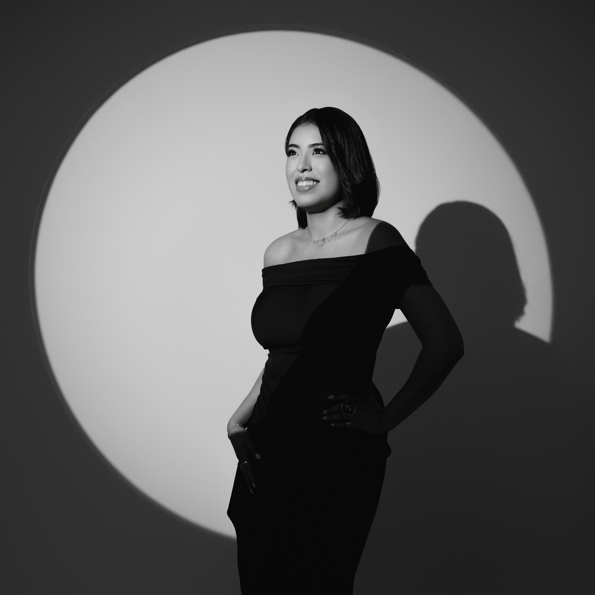 A woman wearing an off-shoulder dress poses against a circular light backdrop in black and white.