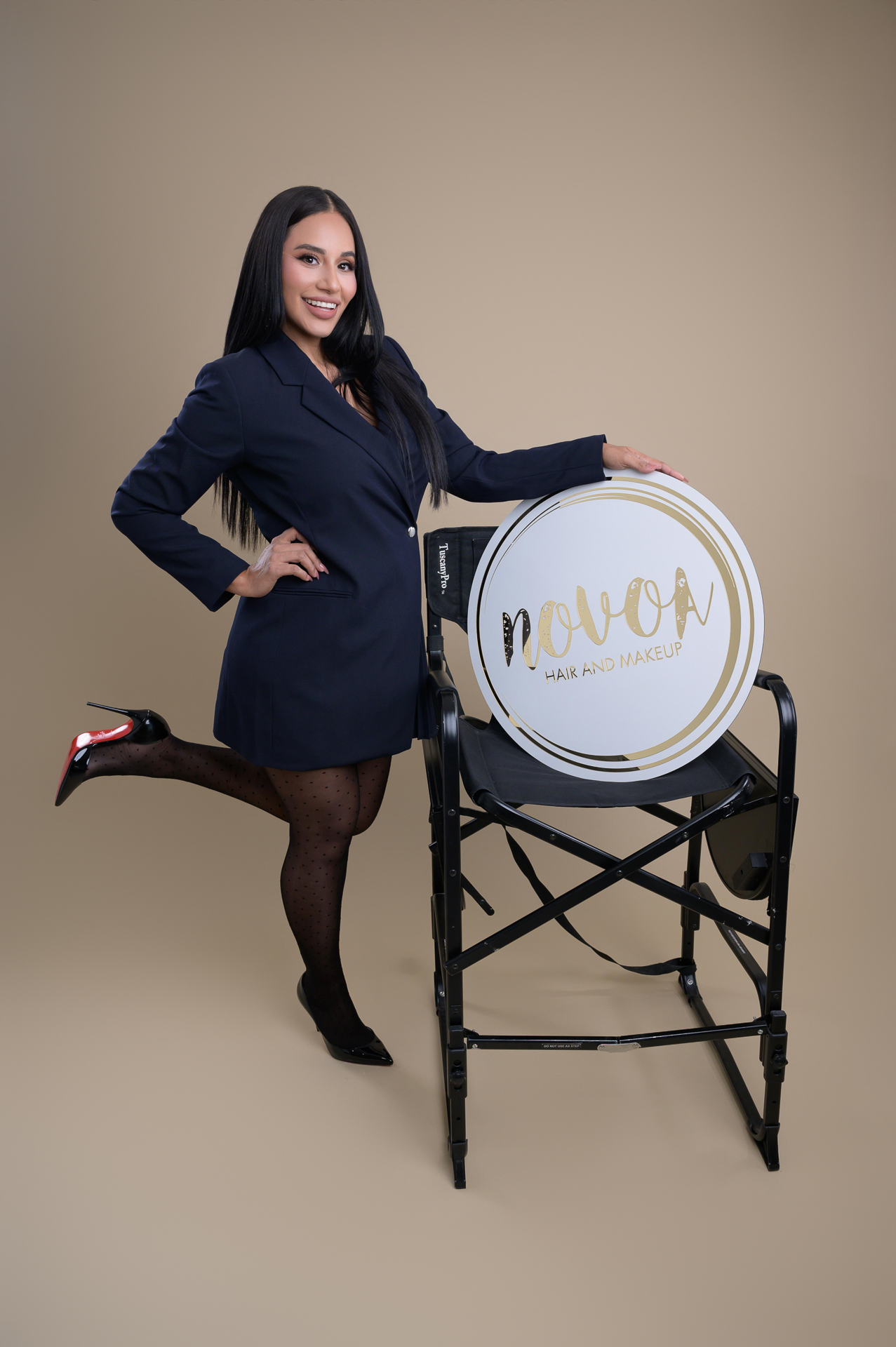 Studio branding portrait of a make up artist in navy blazer and heels stands by a tall chair with Nova Hair and Makeup sign, beige background.