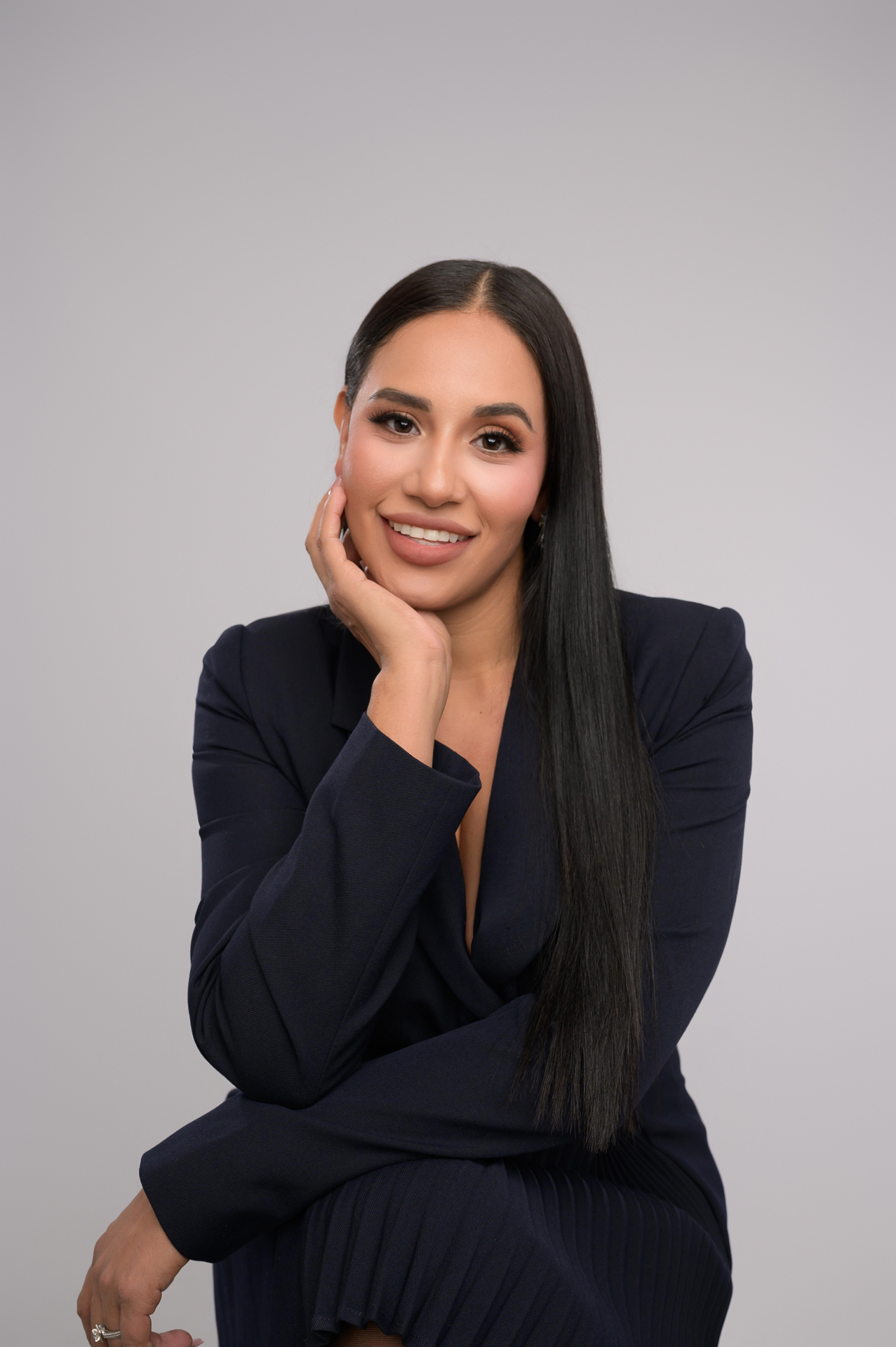 Personal branding photo of a smiling woman in a dark blazer, hand on her cheek, against a light gray background in Gaby Clark Portrait Studio in Los Gatos