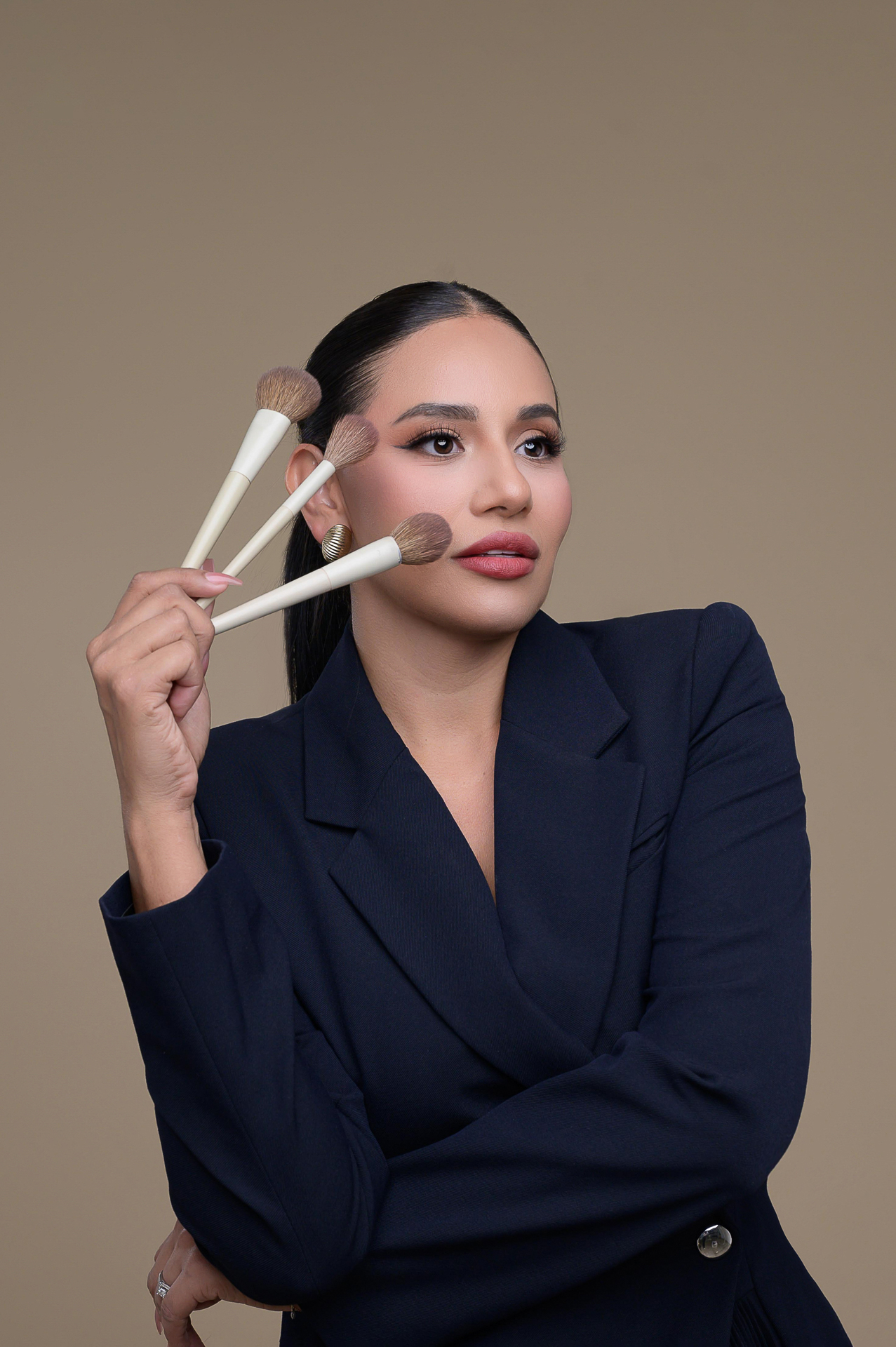 A woman in a dark blazer holding three makeup brushes in her right hand against a neutral background. A woman in a dark blazer holding three makeup brushes in her right hand against a neutral background.