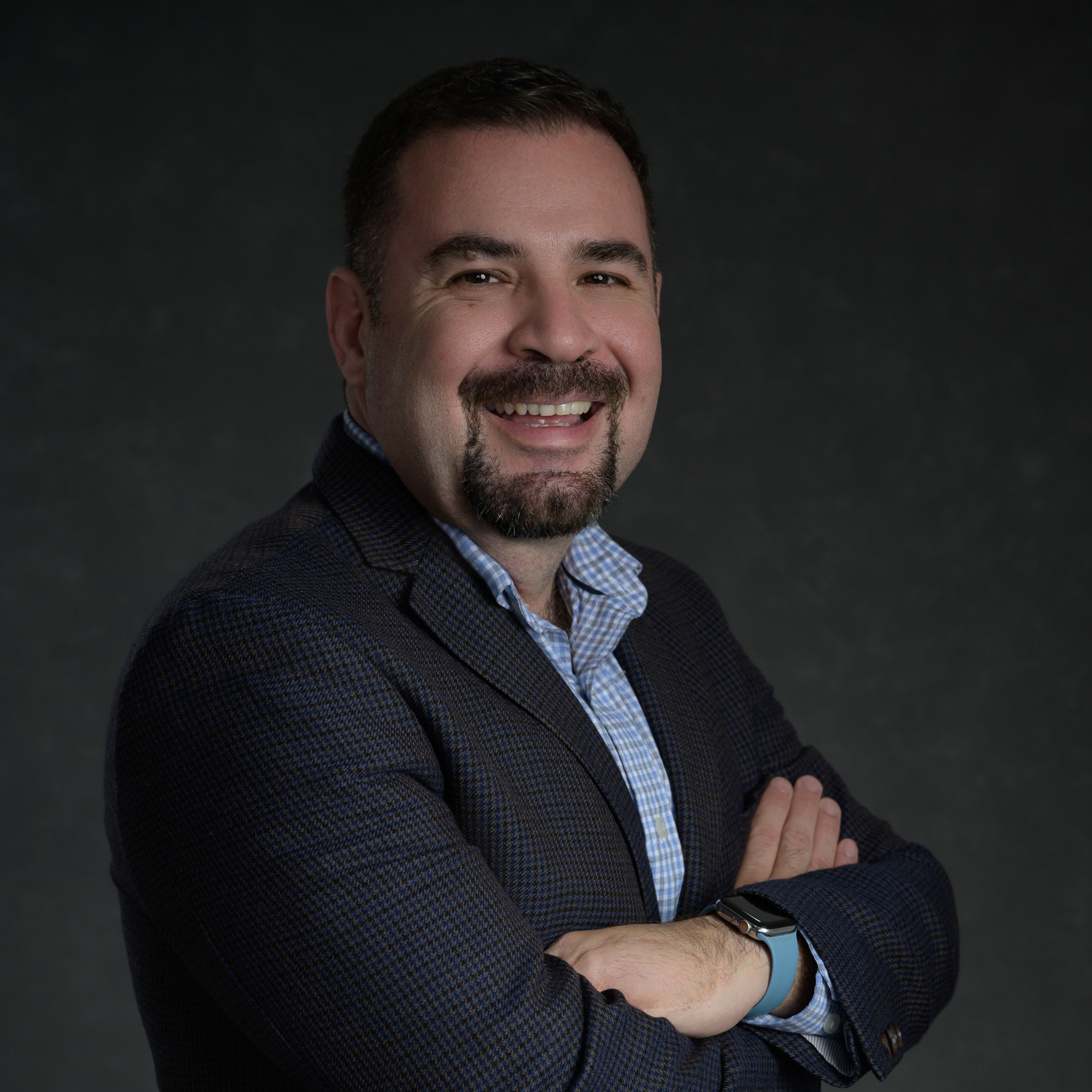 Executive Business Headshots: Man wearing a blazer and blue shirt, smiling with arms crossed against a dark background.