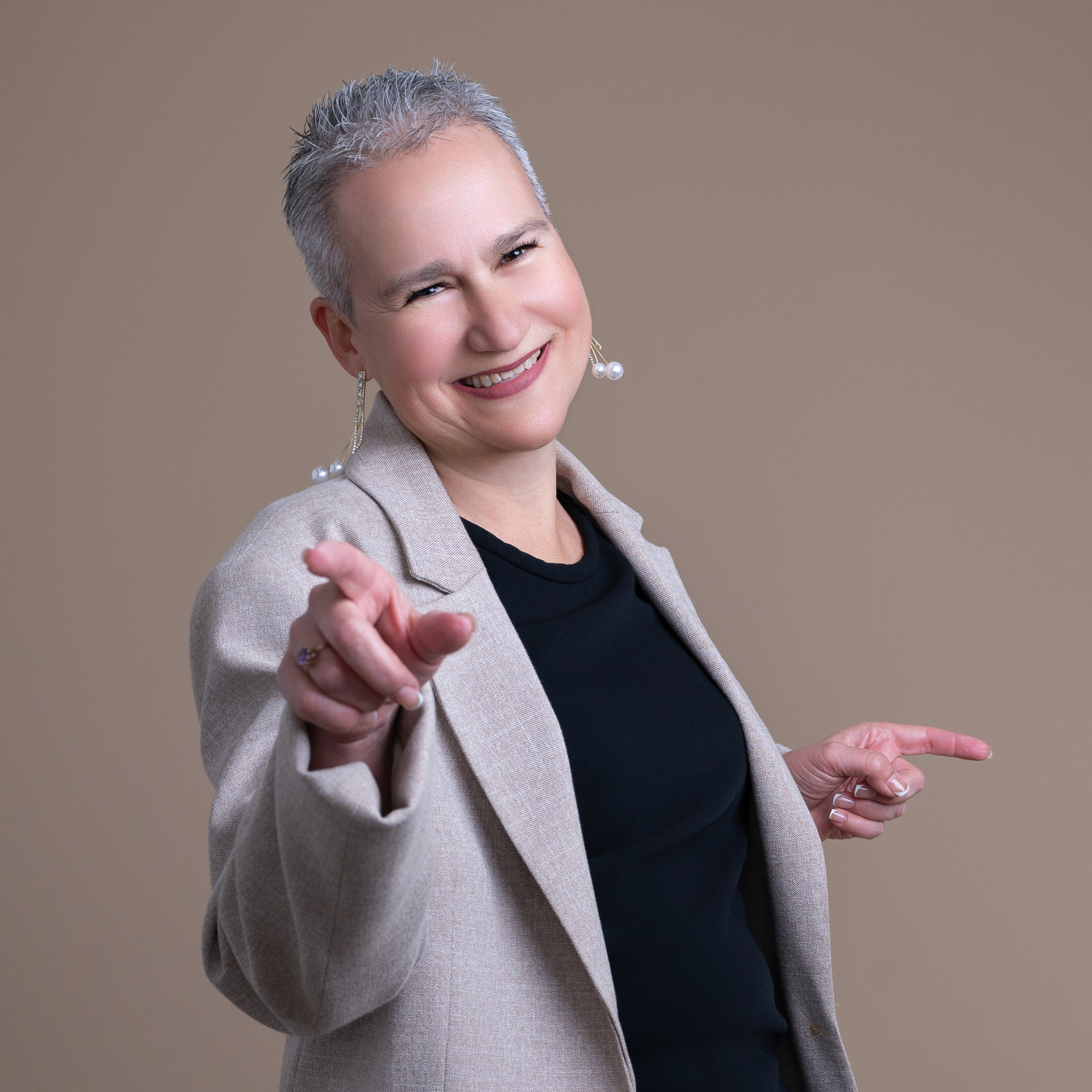 Woman with short gray hair wearing a blazer and large earrings, smiling and pointing with both hands. Happy because she is doing her personal branding session