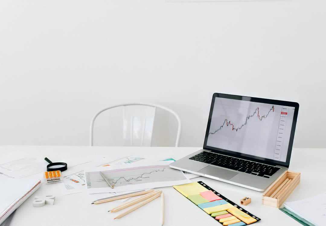 Brand Clarity Ipad Mock up – 7 Personal branding workspace with a laptop chart, papers, calculator, and supplies on a white desk; empty chair in back.