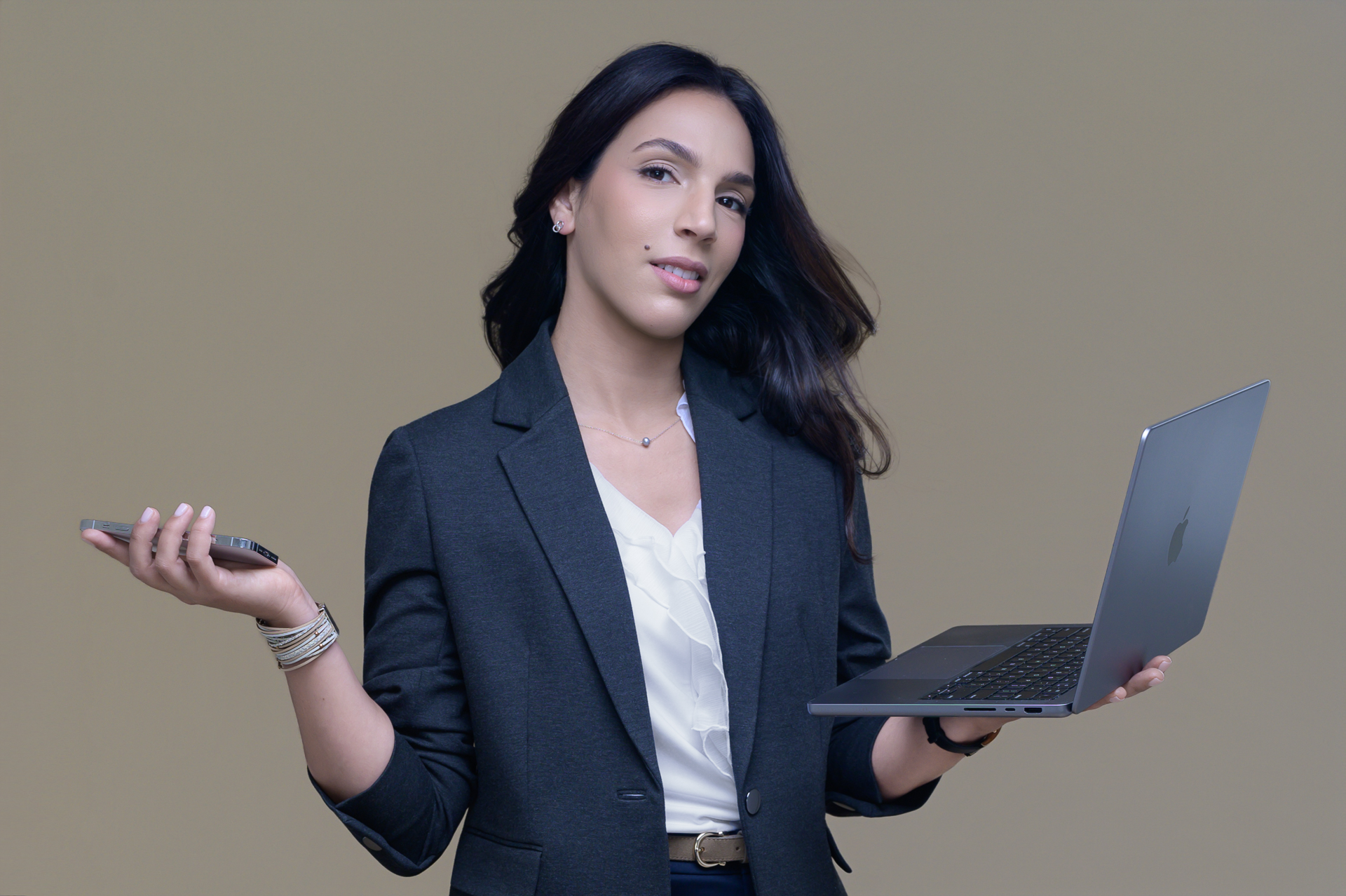 Online Presence Photography: A woman in a business suit holds a phone and laptop, standing with a calm look against a plain beige background.