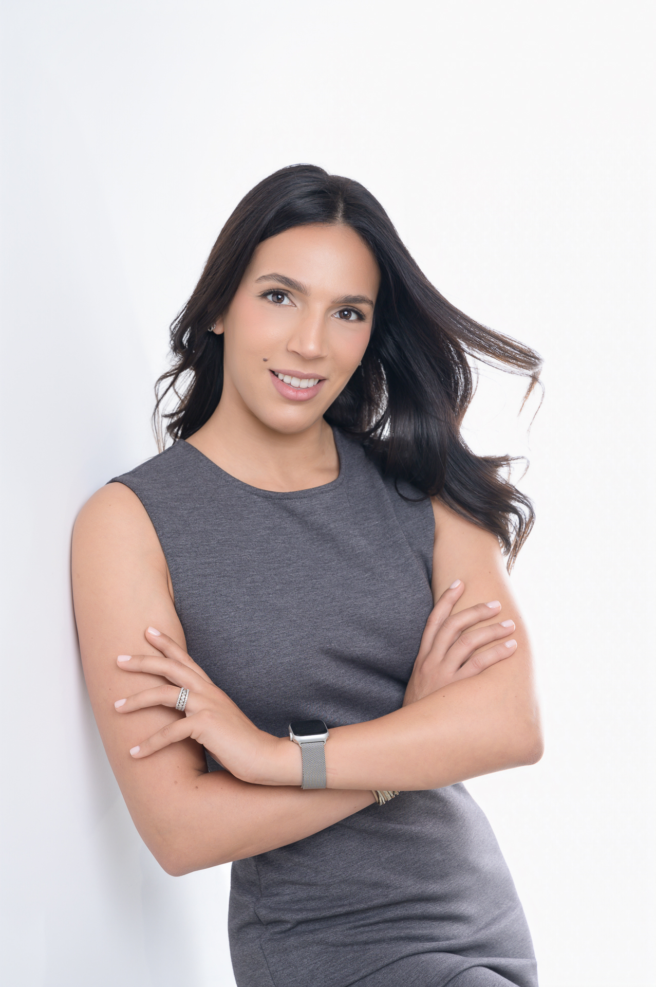 LinkedIn Headshot: A woman with long dark hair wearing a gray dress, posing with arms crossed against a light background.