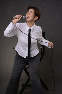 Woman Singing with Microphone Woman in a white shirt and tie singing into a microphone while standing on a chair.