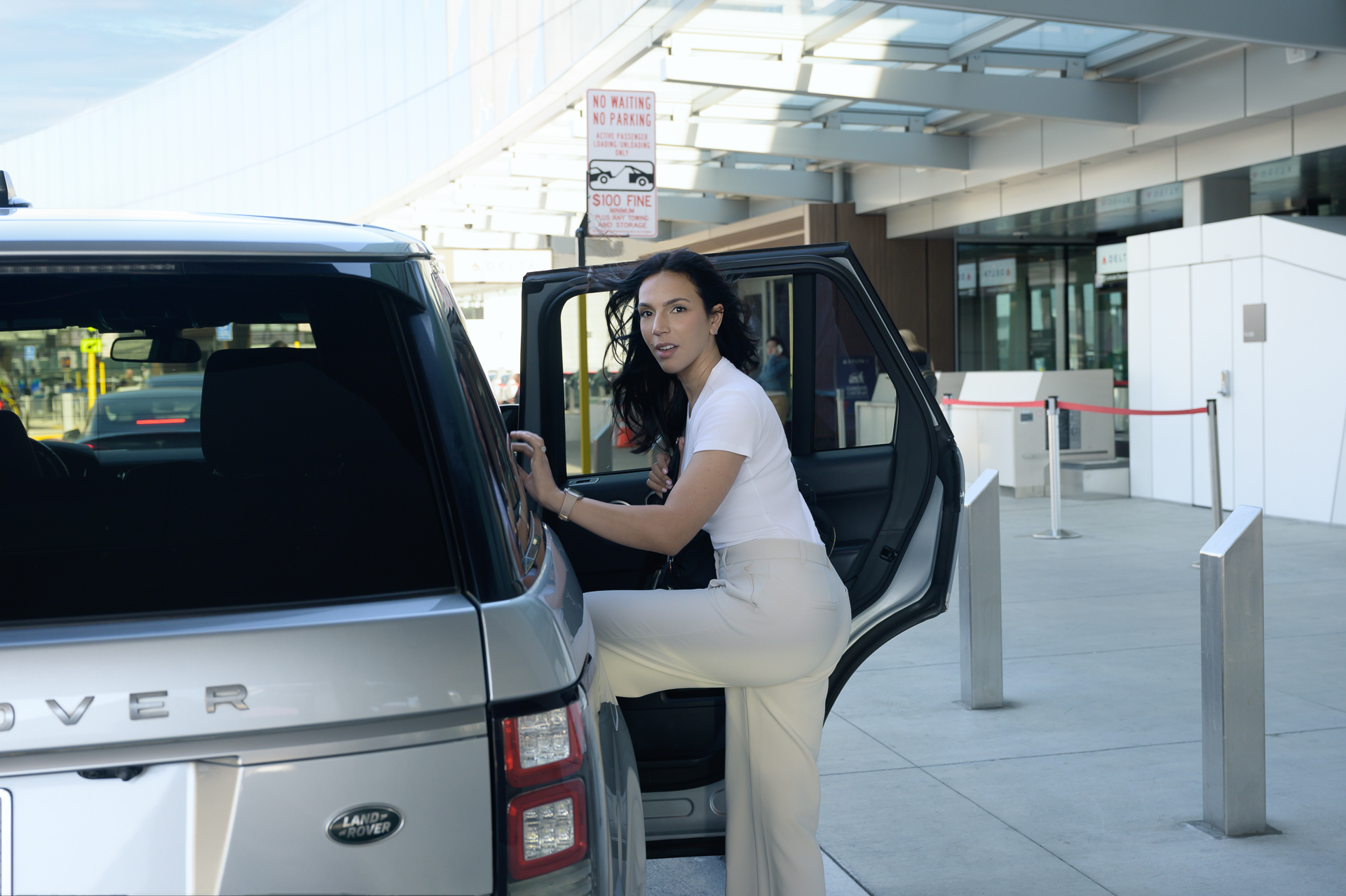 A woman in a white top and beige pants stepping into a silver SUV parked at an airport.