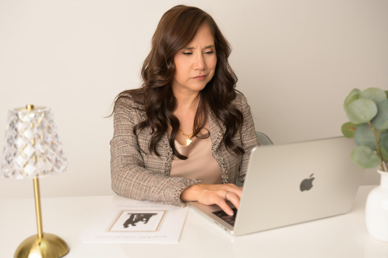 Personal branding in San Francisco Bay Area: Woman with long brown hair works at desk with laptop, lamp, plant, and magazine.