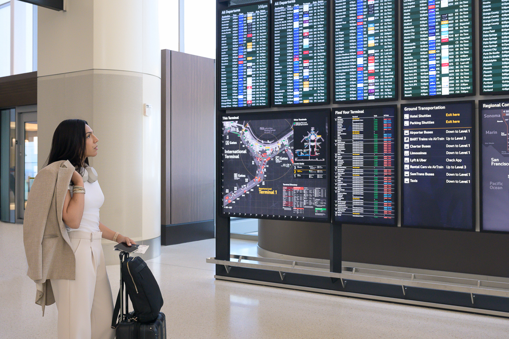 A woman with a suitcase and jacket stands in a bright airport terminal, reading digital flight boards, possibly in San Francisco Bay Area.