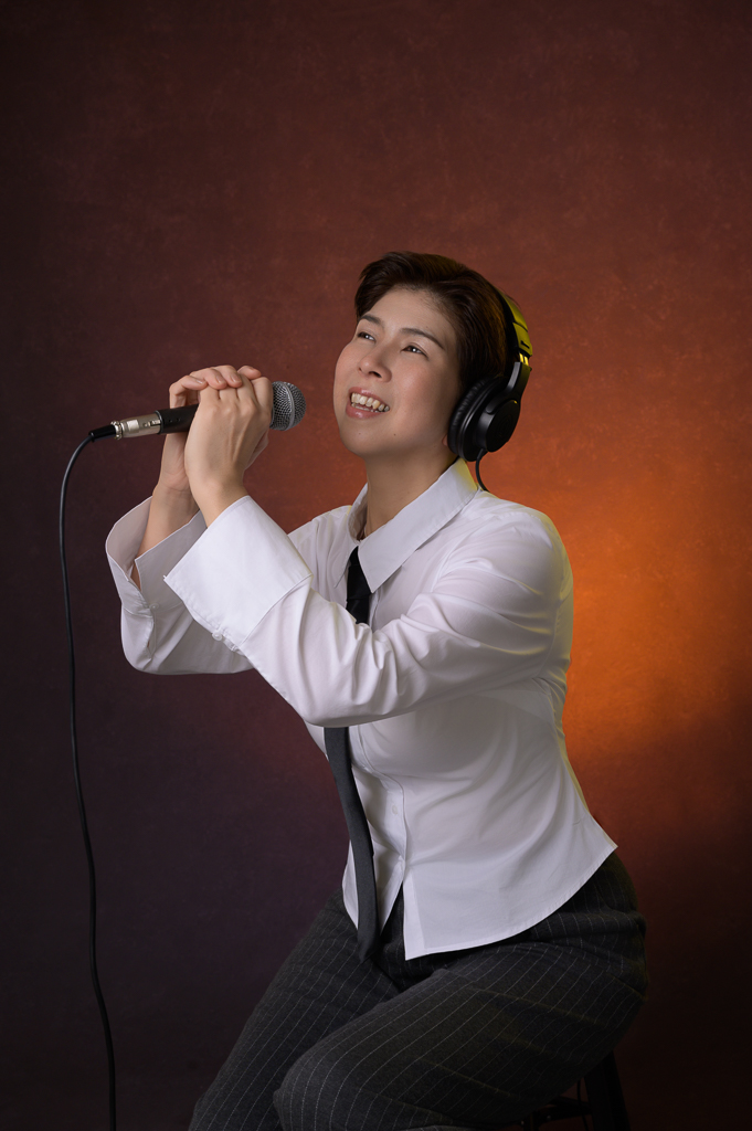 A Mountain View singer in headphones, a white shirt, and black tie sings into a microphone on a stool, lit by warm light against a dark background.
