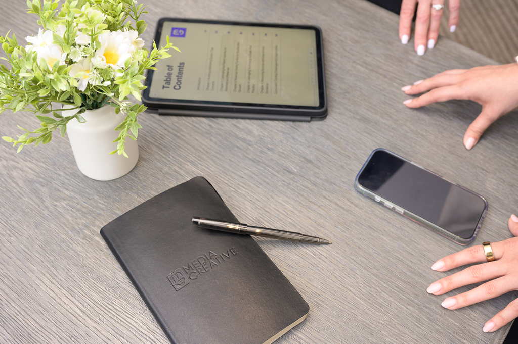 Personal branding workspace with a small vase of flowers, closed Media Creative notebook, phone, tablet showing text, and two ringed hands.