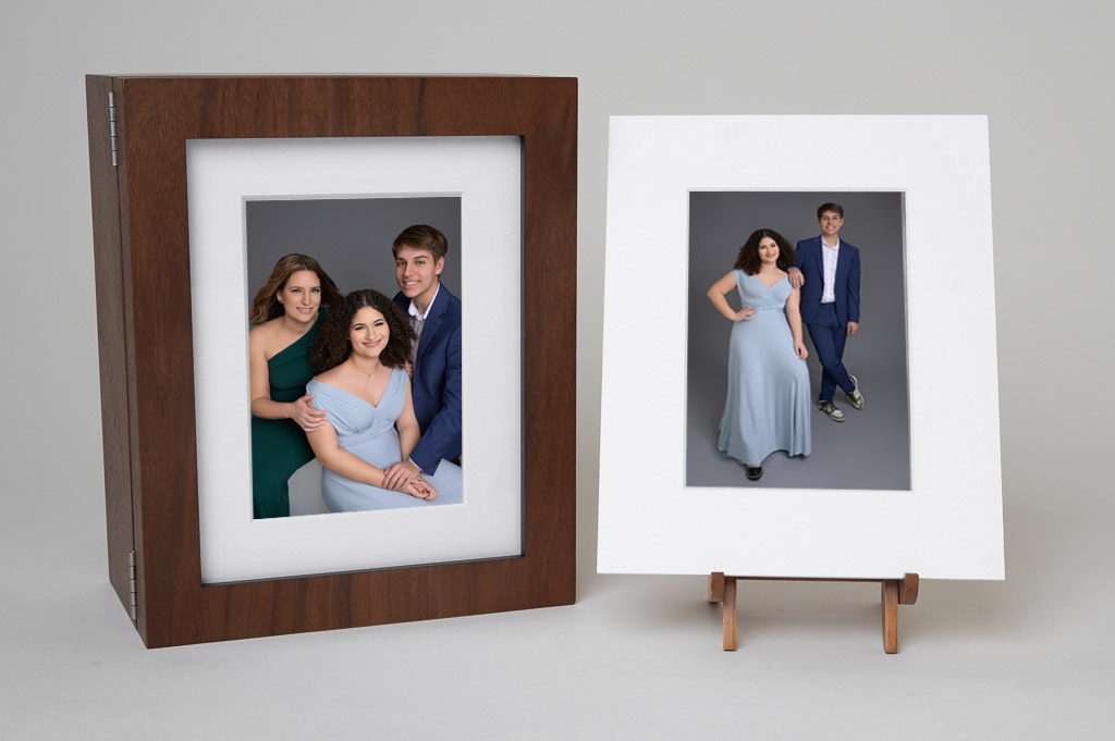 Family portrait photos: Two young adults in formal attire, one seated and smiling, the other standing on a small easel. Plain gray background.