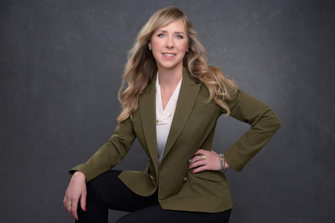 Personal branding photo of a woman with long blonde hair in a green blazer and black pants, seated on a stool against a gray backdrop.
