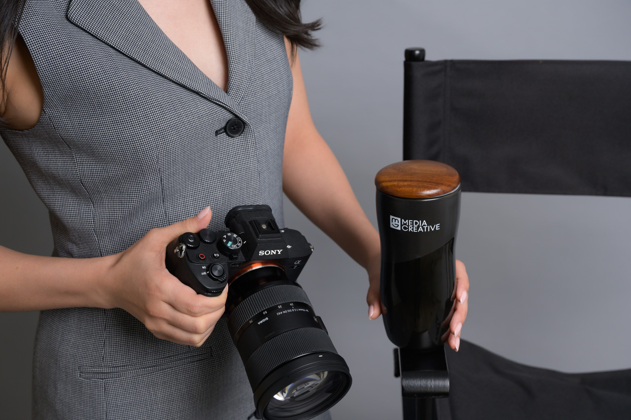 Personal branding photo of a woman in a gray dress holding a Sony camera beside a Media Creative directors chair, gray background.