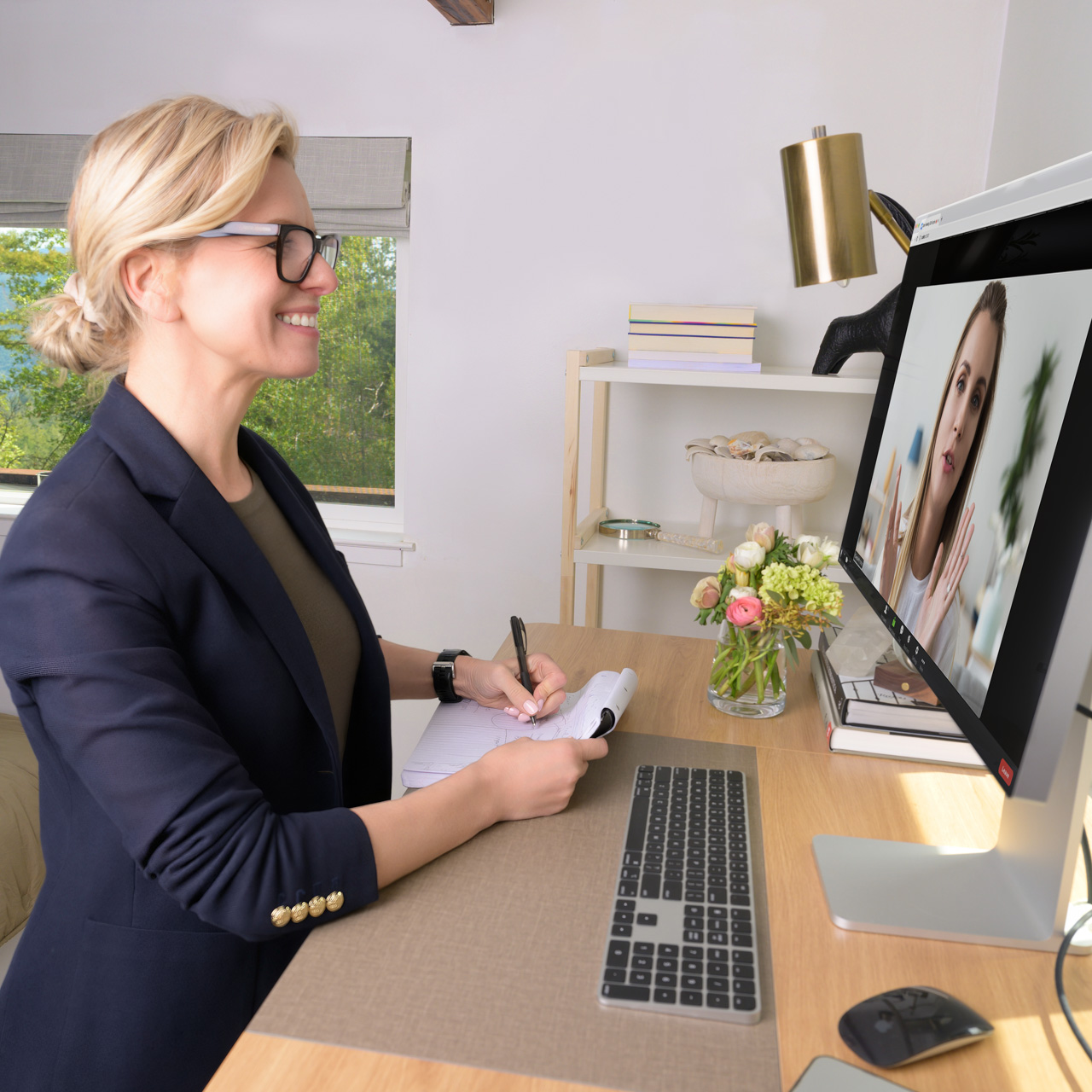 Personal branding session: A woman in glasses and a blazer takes notes at her desk on a video call, with flowers and office supplies nearby.